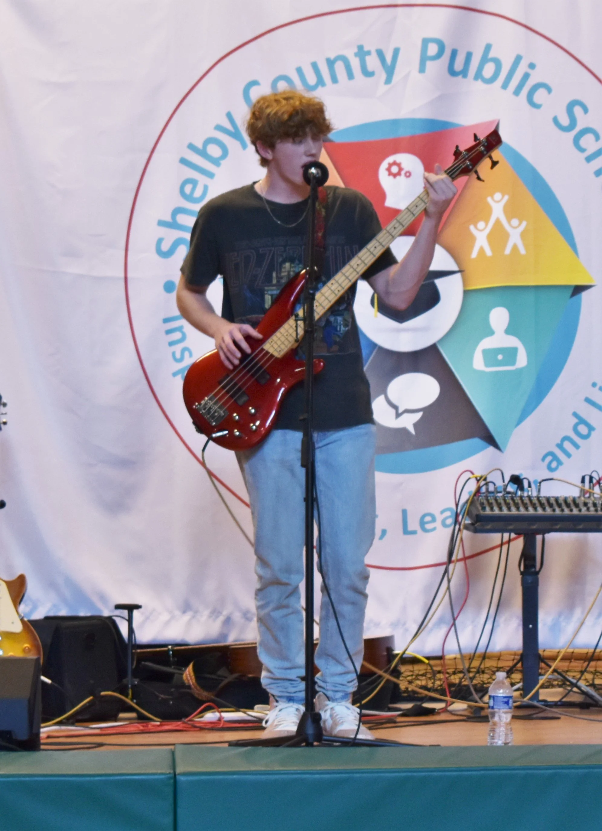 Young male musician with curly hair playing a red bass guitar on stage, singing into a microphone, at an event with a large colorful banner in the background.