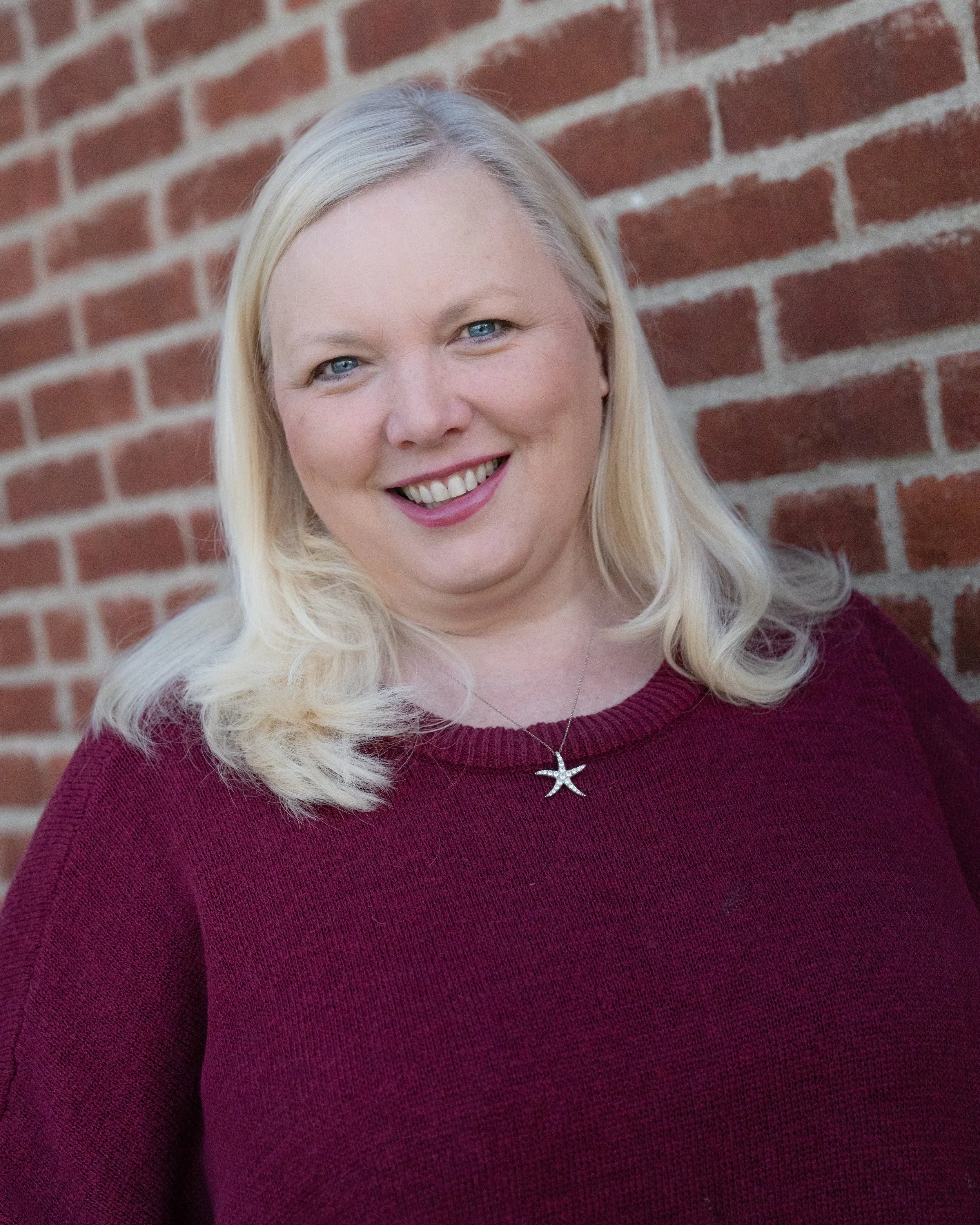 A woman with blonde hair and blue eyes smiling, wearing a maroon sweater and a starfish necklace, standing against a brick wall.