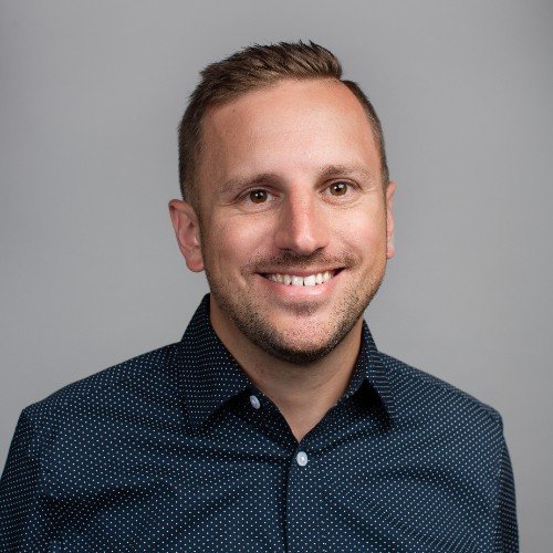 A smiling man with short brown hair, wearing a dark blue button-up shirt with small white dots, against a plain gray background.