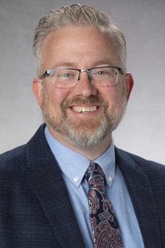 A smiling man with glasses, a beard, and gray hair, wearing a dark blazer, light blue shirt, and paisley tie, against a gray background.