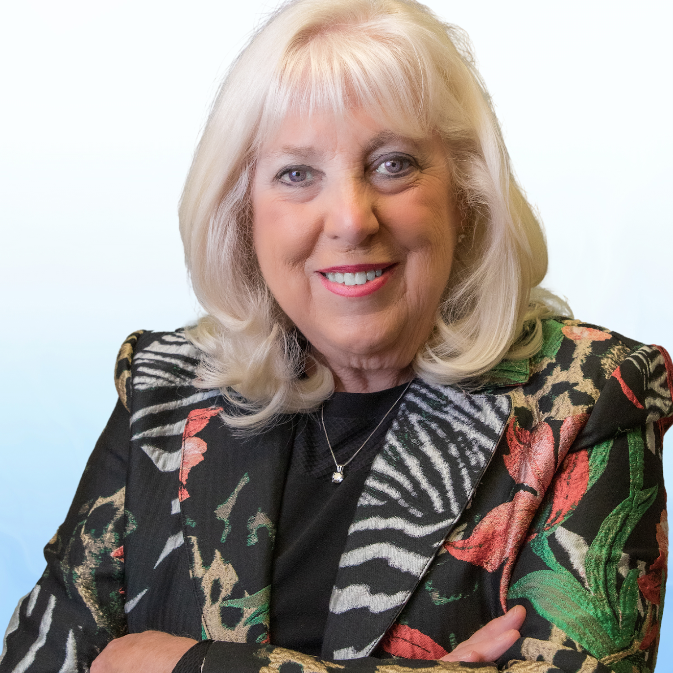 A smiling elderly woman with blonde hair and blue eyes, wearing a colorful floral blazer and a black top, with a silver necklace, against a light background.