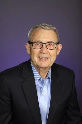 Portrait of a middle-aged man with glasses, wearing a dark suit jacket and blue striped shirt, smiling against a dark background.
