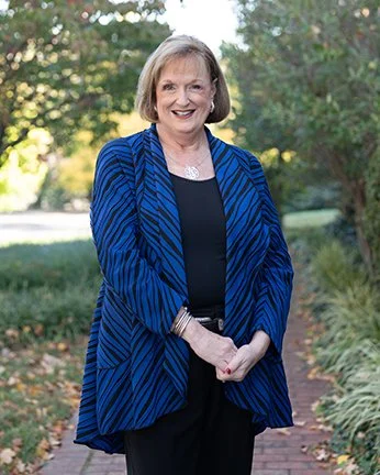 A smiling woman in a blue patterned blazer standing outdoors on a brick path with greenery and trees in the background.