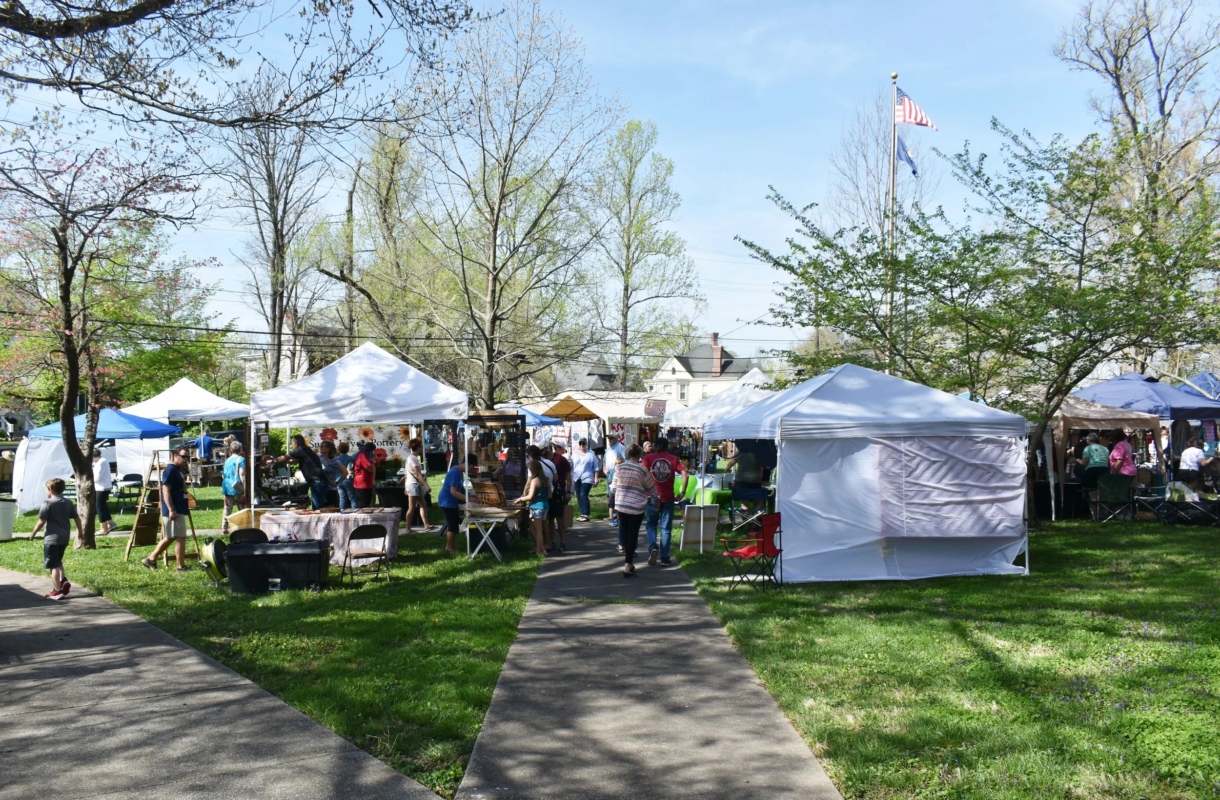A lively outdoor market with white tents and people shopping on a clear spring day, surrounded by trees with some blooming.