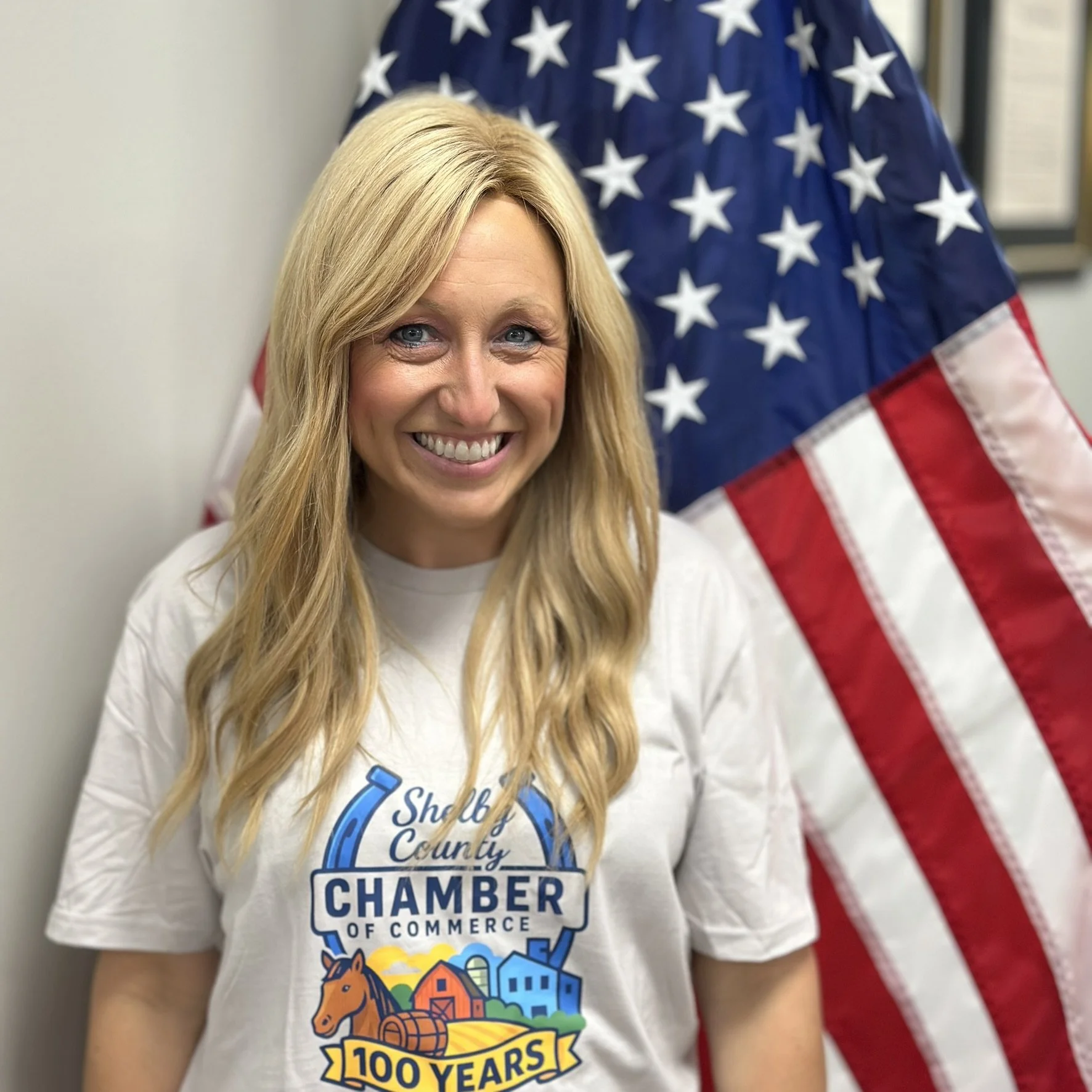 A smiling blonde woman with long wavy hair wearing a white T-shirt with the logo of Shelley County Chamber of Commerce celebrating 100 years. Behind her is a large American flag.