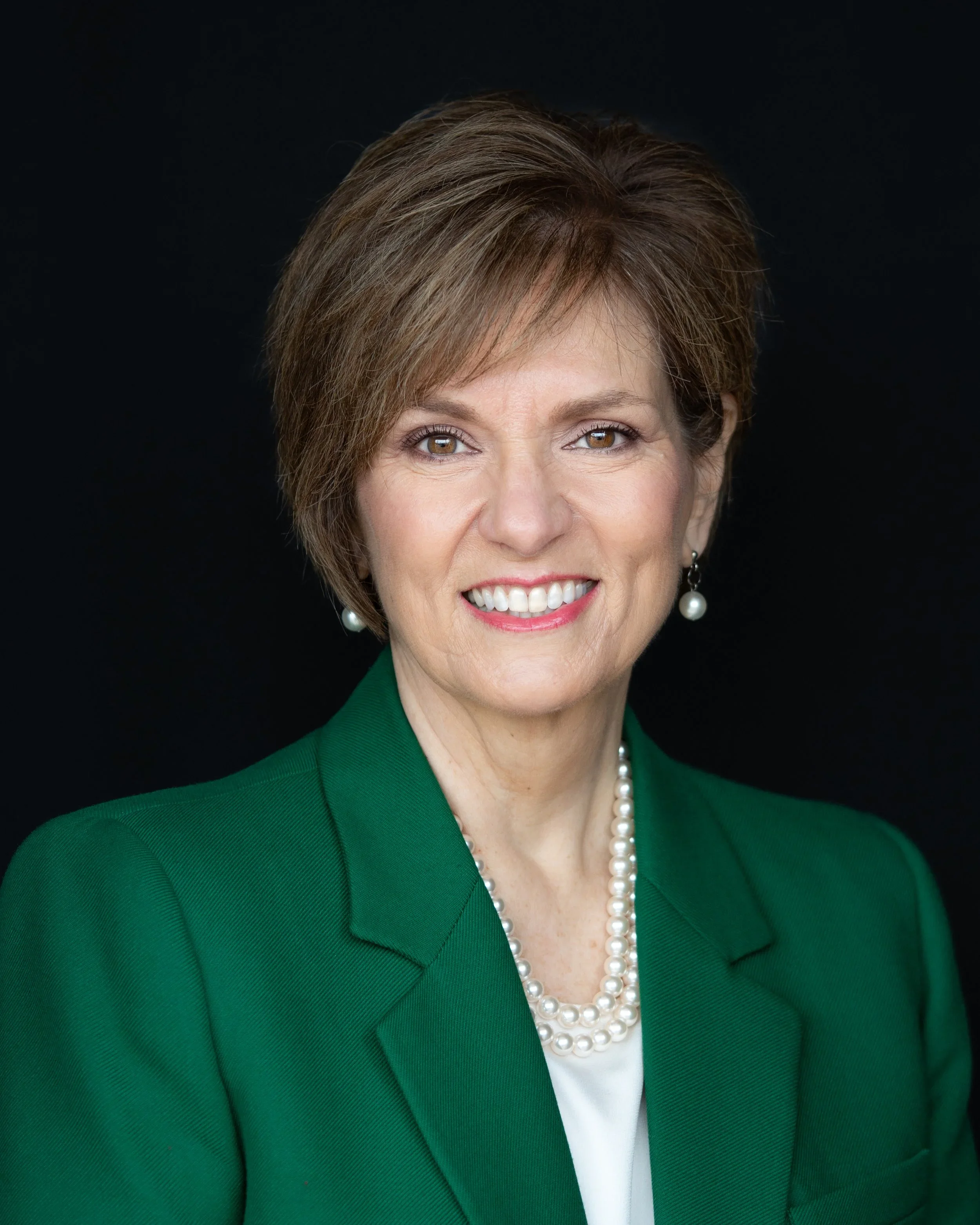 A woman with short brown hair, wearing a green blazer, white shirt, and pearl necklace, smiling against a dark background.