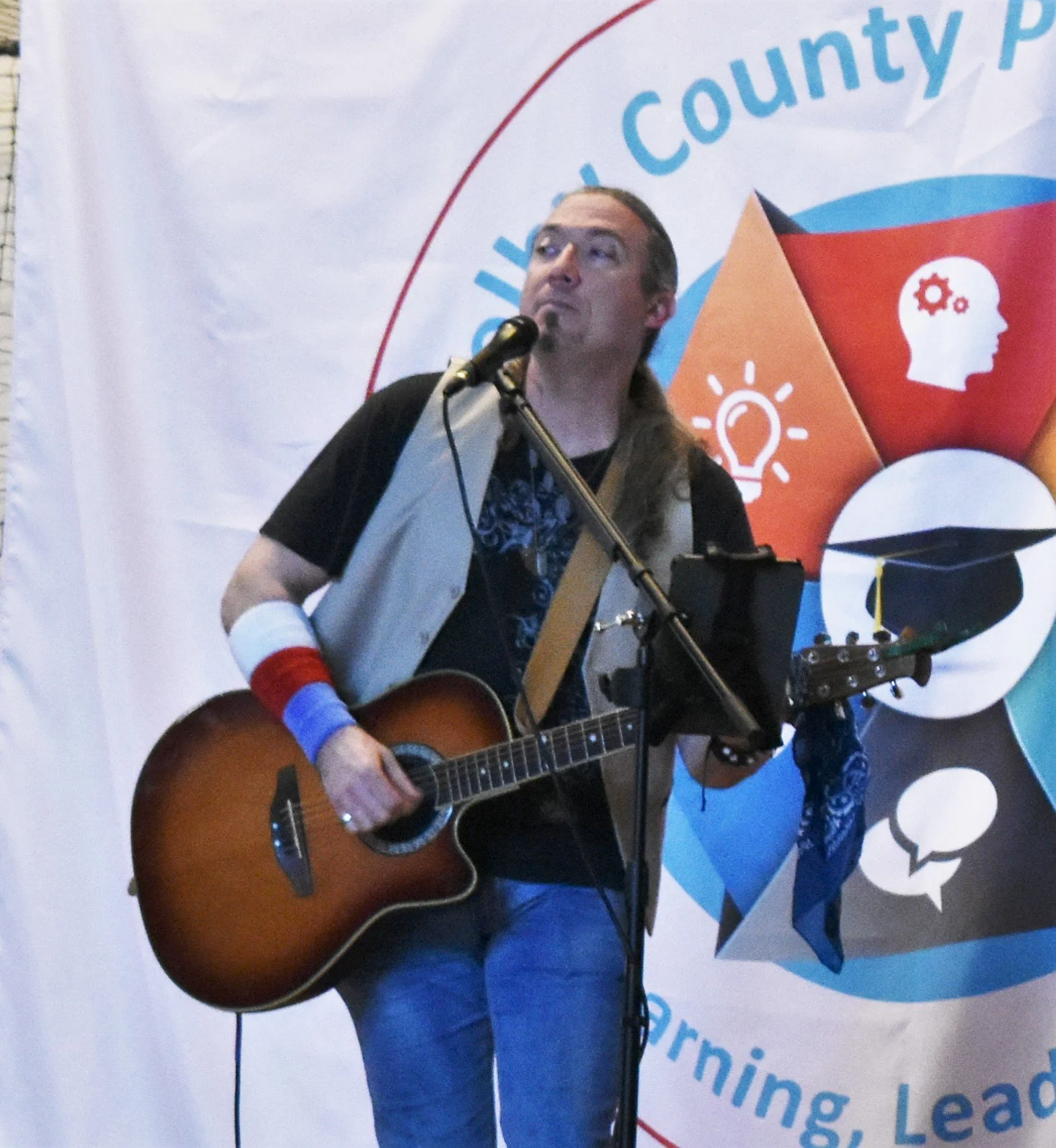 A man with long hair playing an acoustic guitar and singing into a microphone at an event with a colorful backdrop that has a brain, light bulb, graduation cap, and speech bubble icons and text related to learning and leadership.