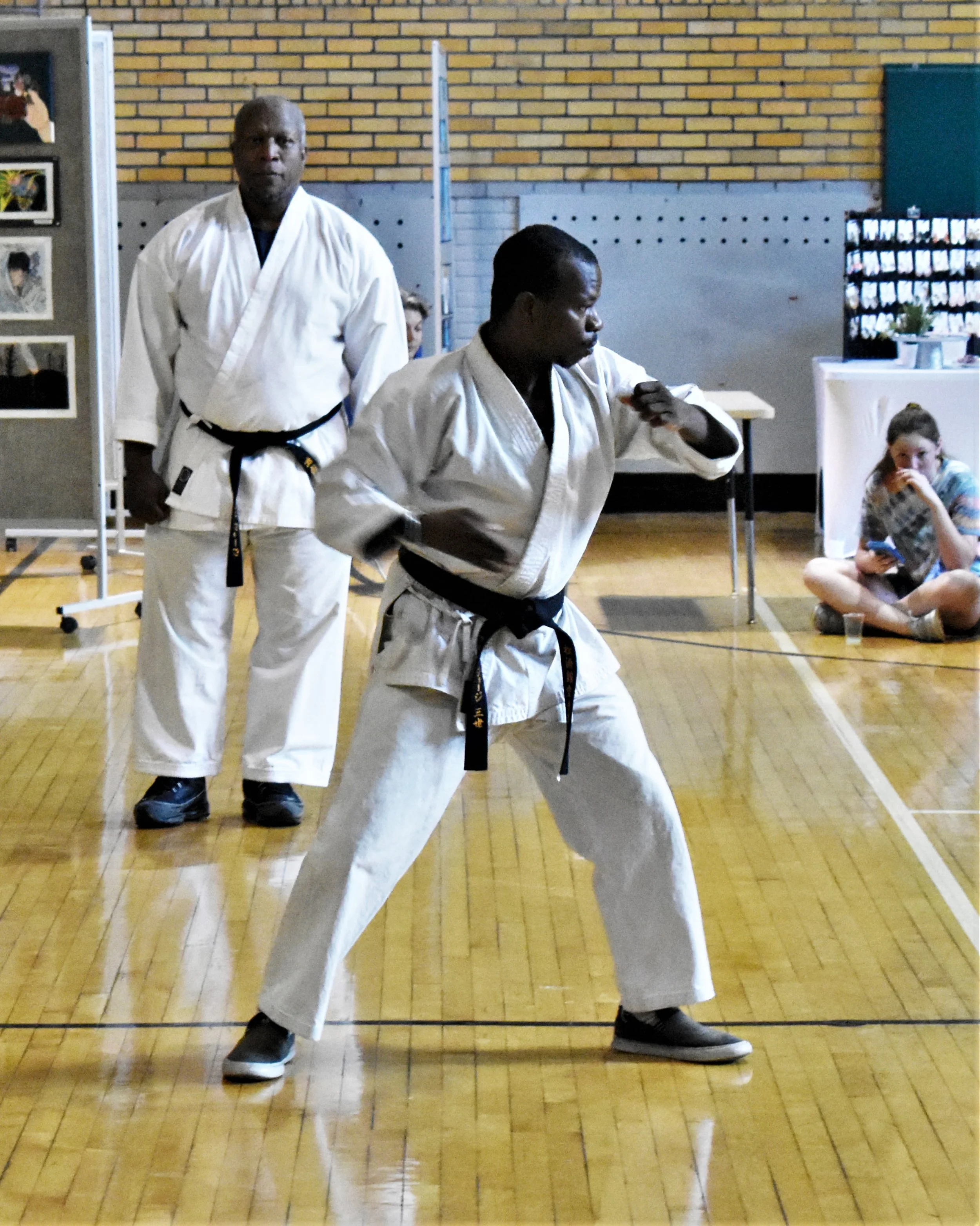 A person practicing karate in a gymnasium with a black belt, performing a stance or move, while an instructor in a white karate gi observes. In the background, a woman is sitting on the floor reading a book and a kid is sitting next to her.