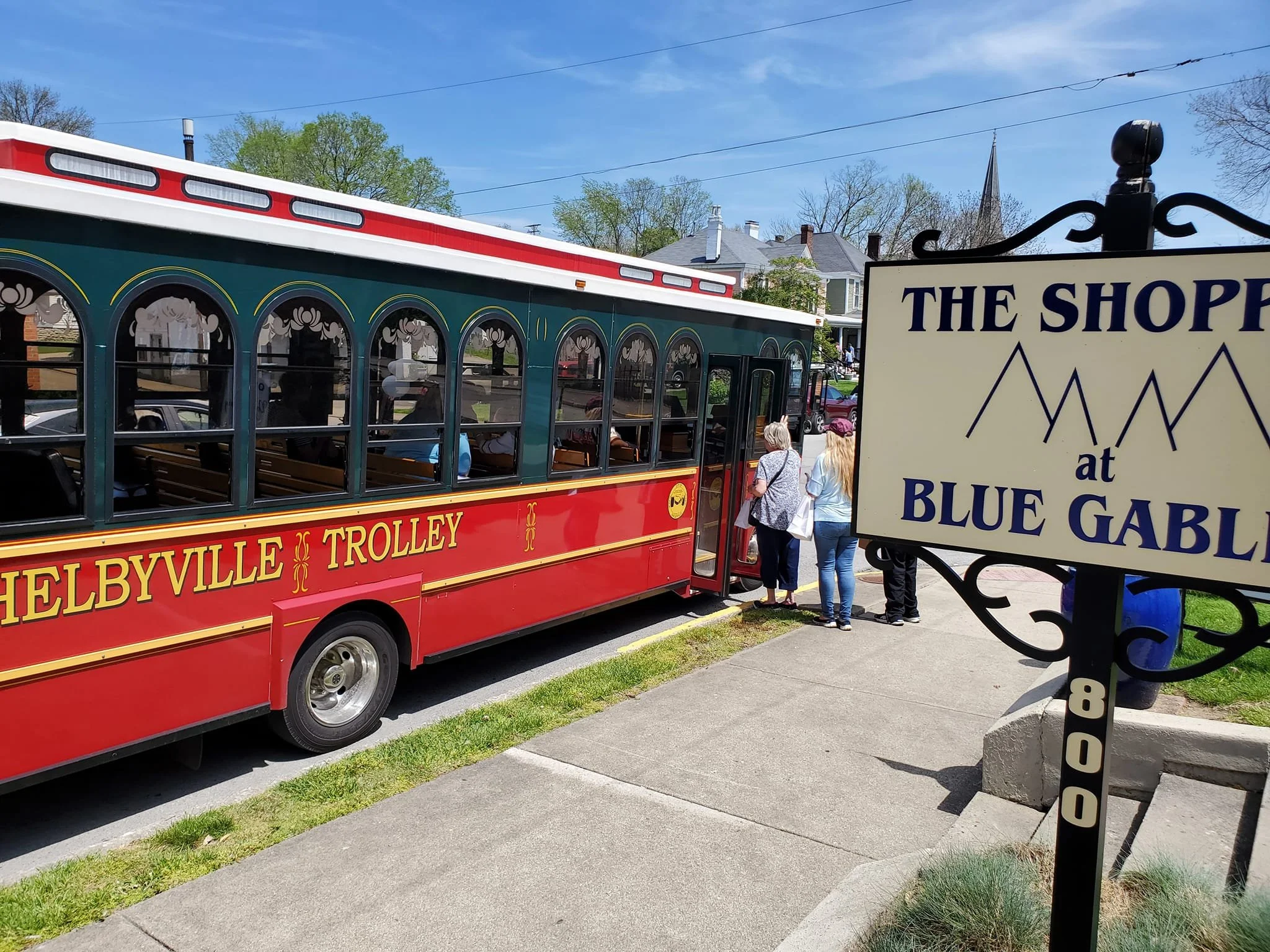 Red trolley bus labeled 'Shelbyville Trolley' parked at the stop with a group of people boarding, next to a sign that reads 'The Shops at Blue Gables'.
