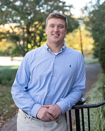 A young man smiling outdoors on a sunny day, wearing a light blue dress shirt and khakis, standing near a black metal fence with trees and a pathway in the background.