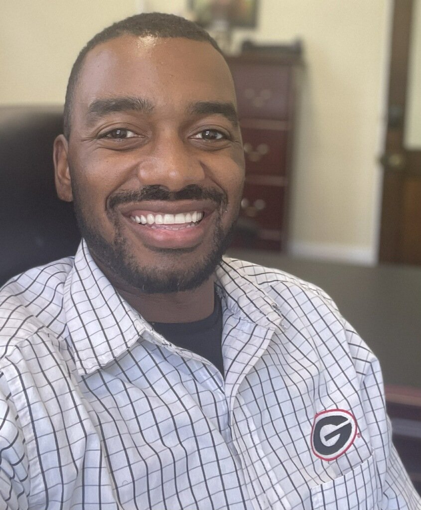 A smiling man with a beard and short hair wearing a checkered shirt with a Georgia Bulldogs logo, sitting in an office chair with a dresser and open door in the background.