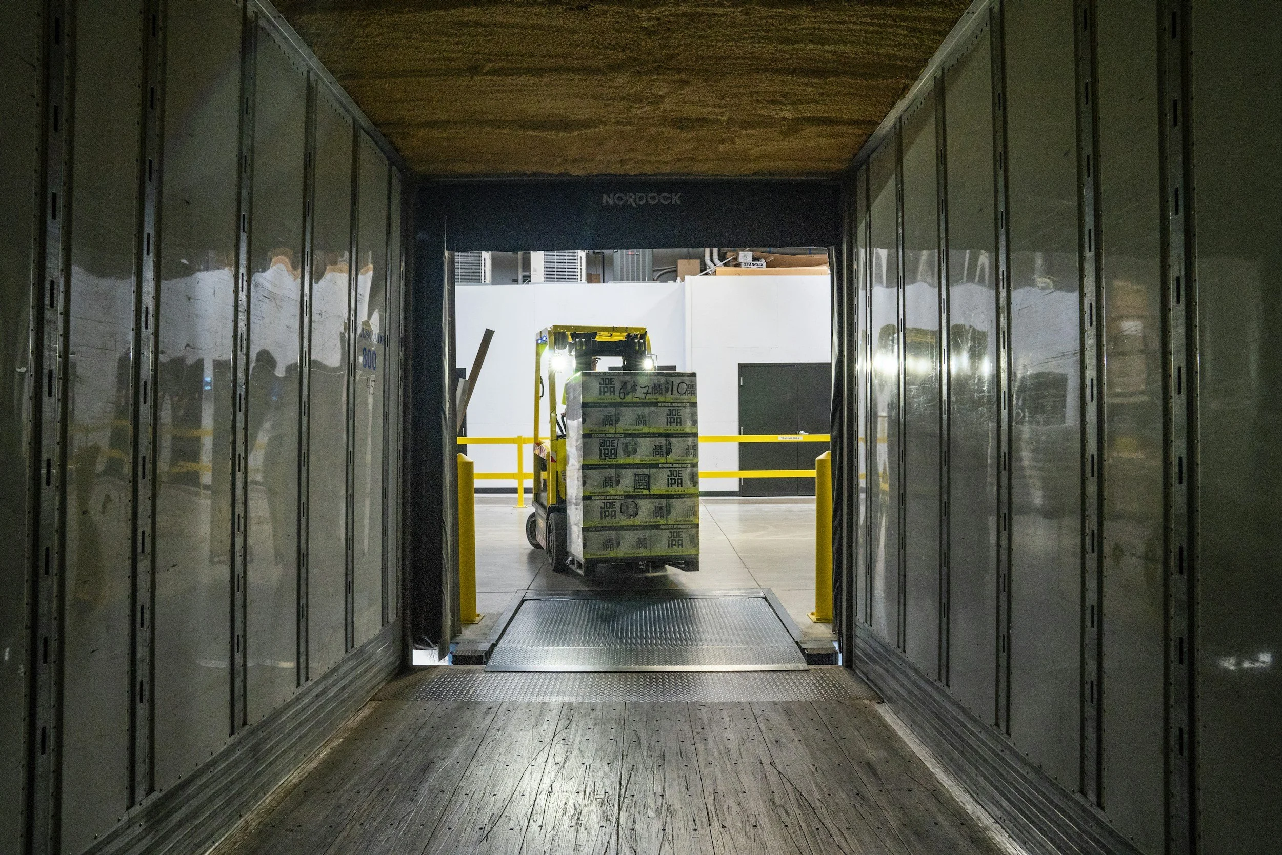 View from an indoor loading dock or corridor leading to an outdoor storage area with a pallet jack carrying boxes of beer. Bright lights illuminate the area with yellow safety barriers.