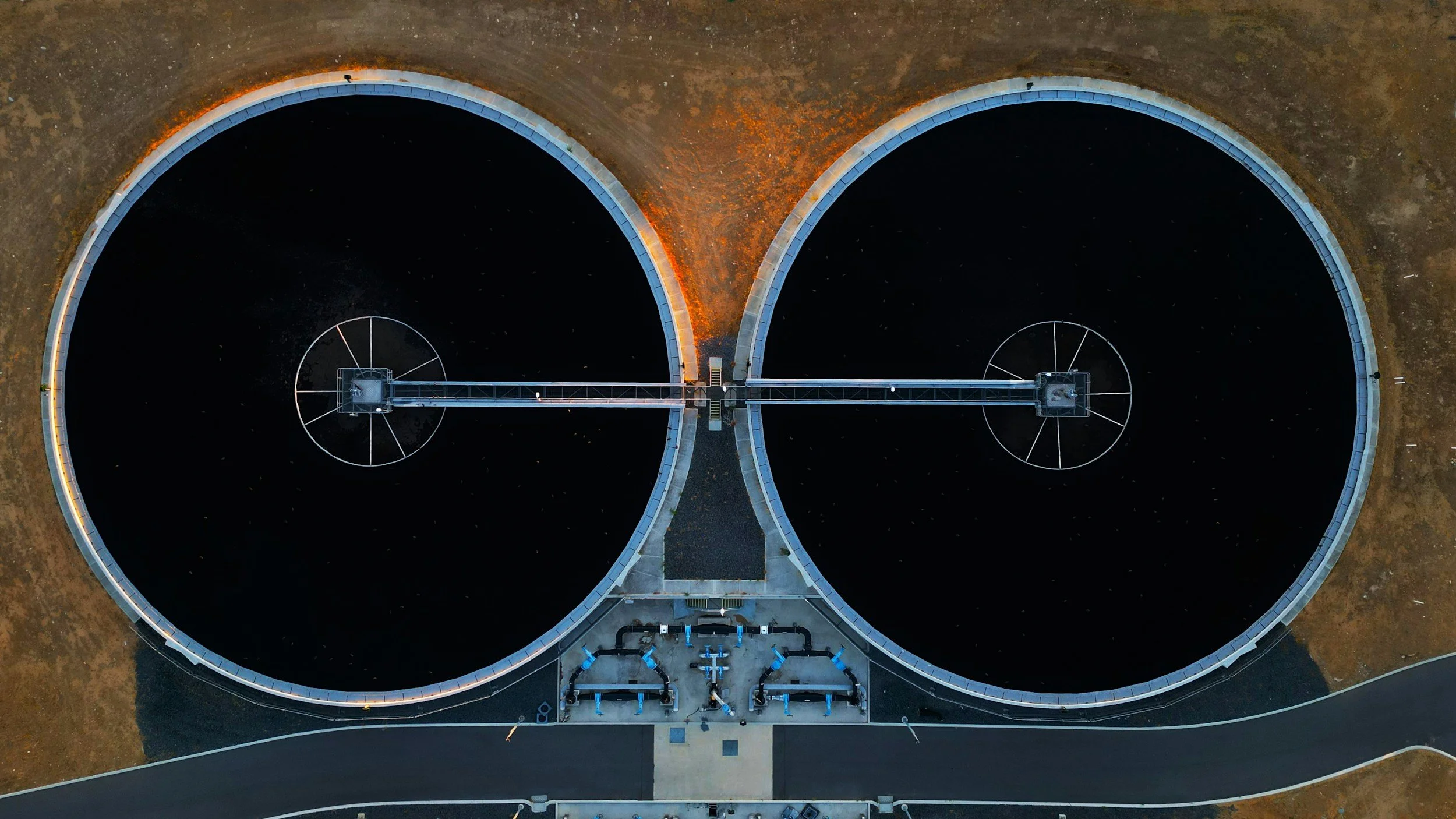 An aerial view of two large circular water reservoirs with connecting pipes and surrounding infrastructure, illuminated at night.