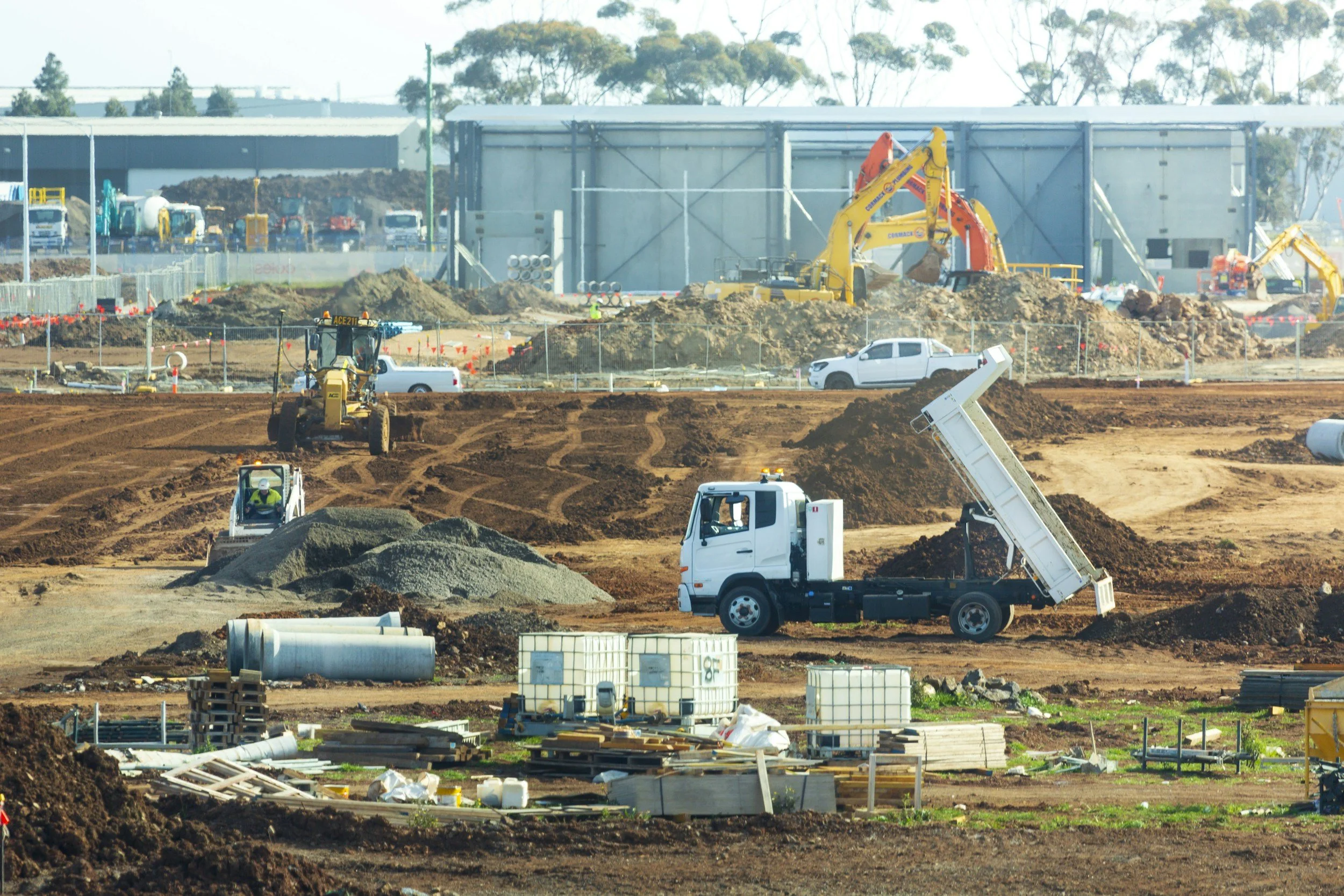 Construction site with multiple construction vehicles, including dump trucks, excavators, and bulldozers, working on dirt and soil. Piles of gravel and building materials are scattered around the area.