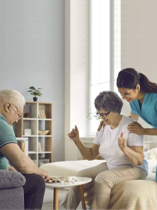 Three women playing checkers or chess at a small round table in a bright, home setting, with a bookshelf and large windows in the background.