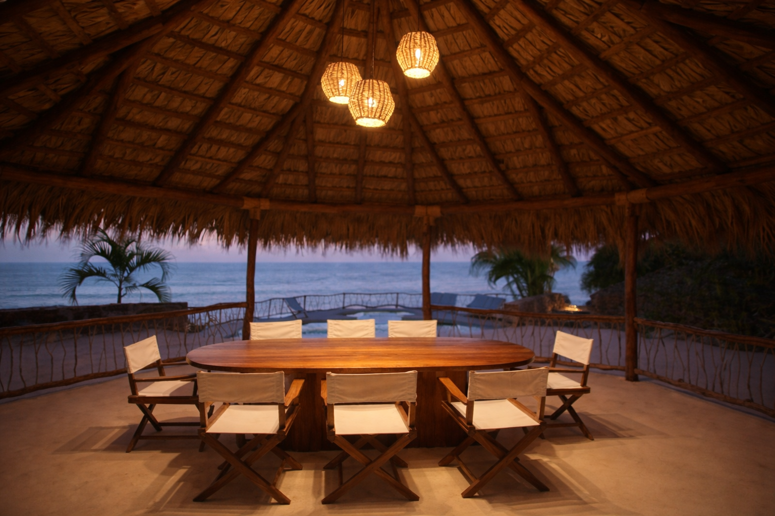An open-air palapa dining space at ZAIA, a luxury private villa in Zihuatanejo, Mexico. Handcrafted wood furnishings and warm pendant lighting create an intimate setting for shared meals overlooking the Pacific coastline at sunset.