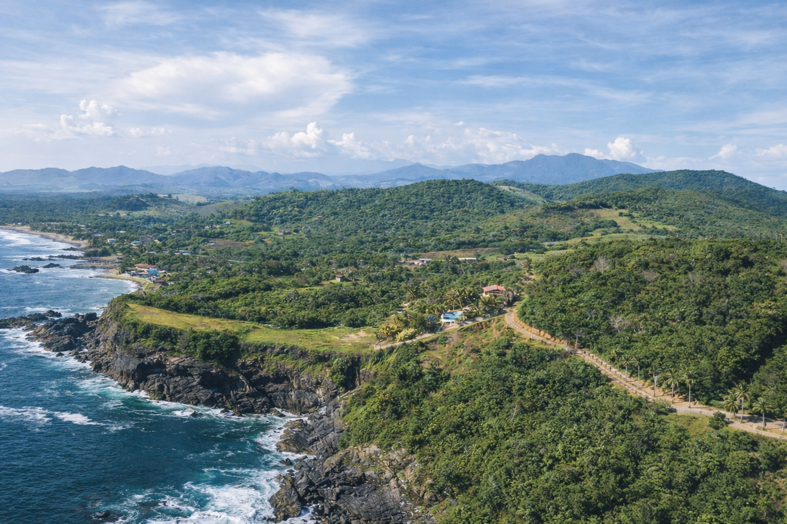 An aerial view of the rugged Pacific coastline near ZAIA, a private cliffside villa in Zihuatanejo. Rolling green hills, rocky cliffs, and expansive ocean views define the natural landscape surrounding the property.