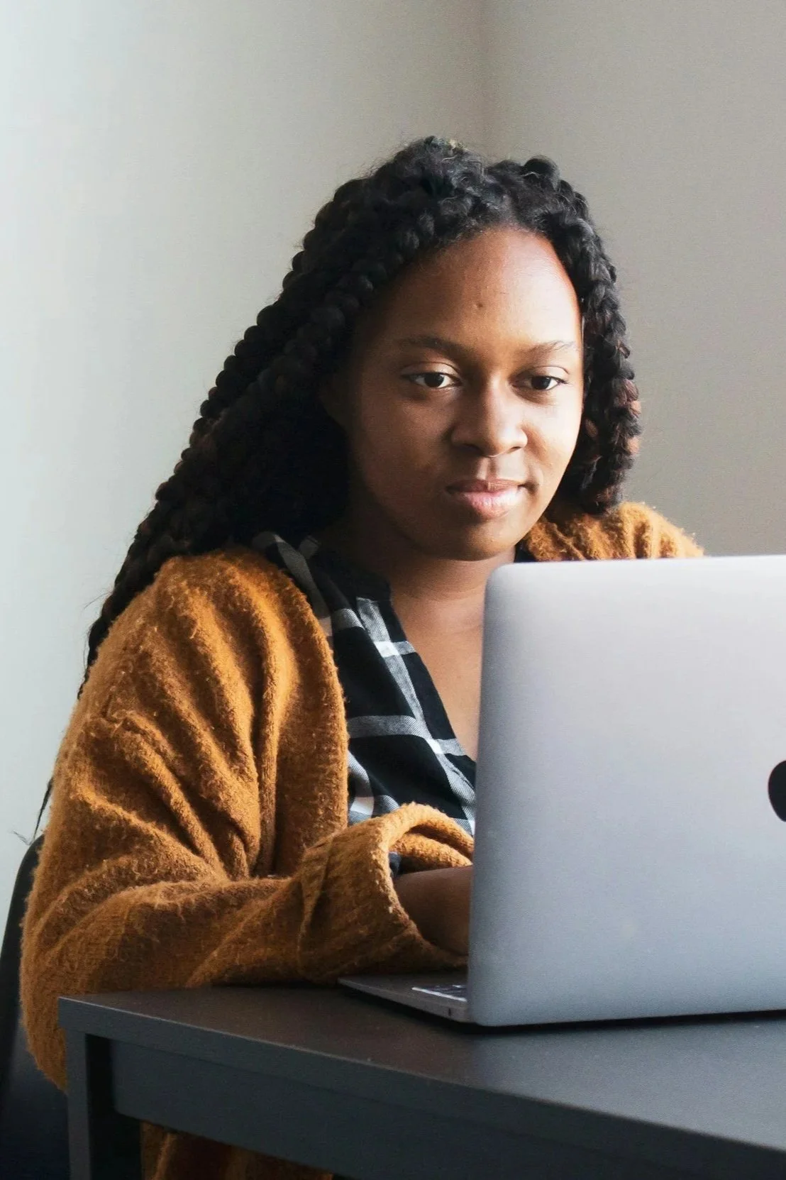 A woman with braided hair sitting at a table using a laptop, looking at the camera.