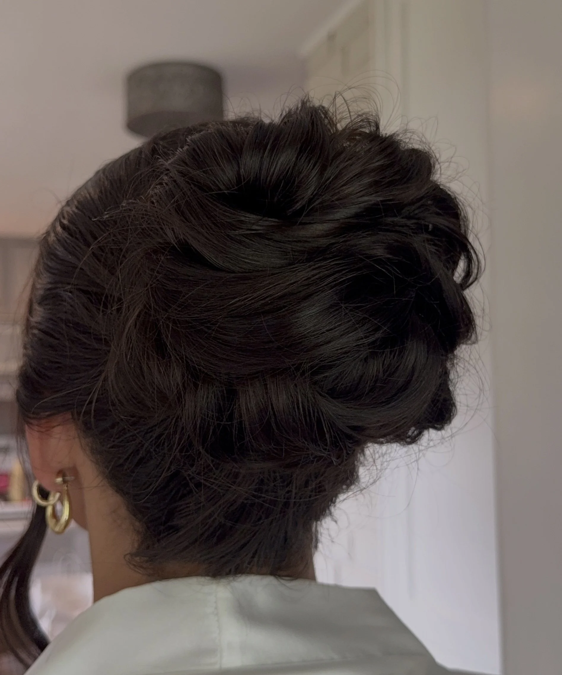Close-up of a person's dark brown curly hair styled in an elegant updo, with a side view of the person's head showing the hairstyle.