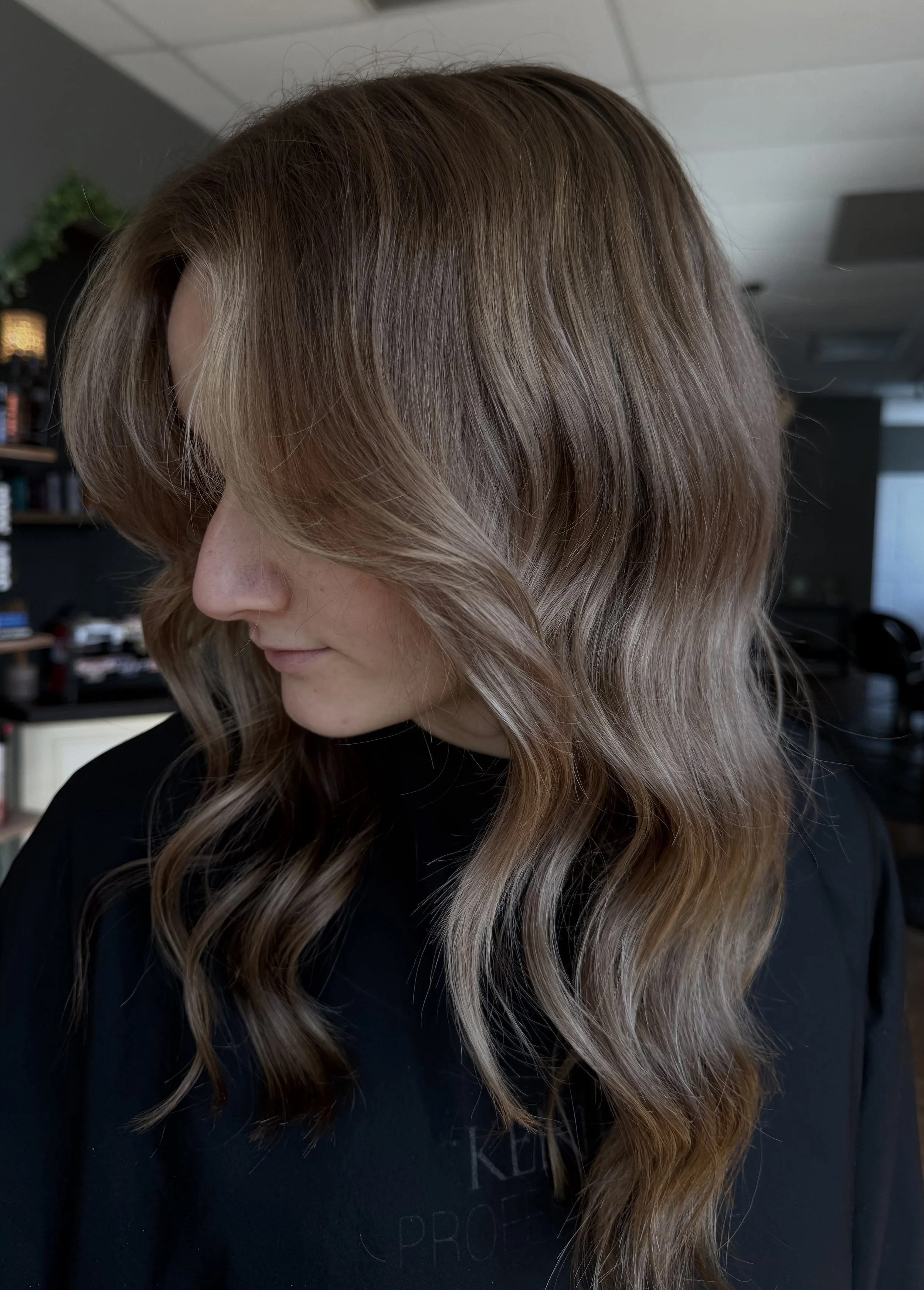 A woman with long, wavy, light brown hair that has subtle highlights, looking downward, in a salon or indoor setting.