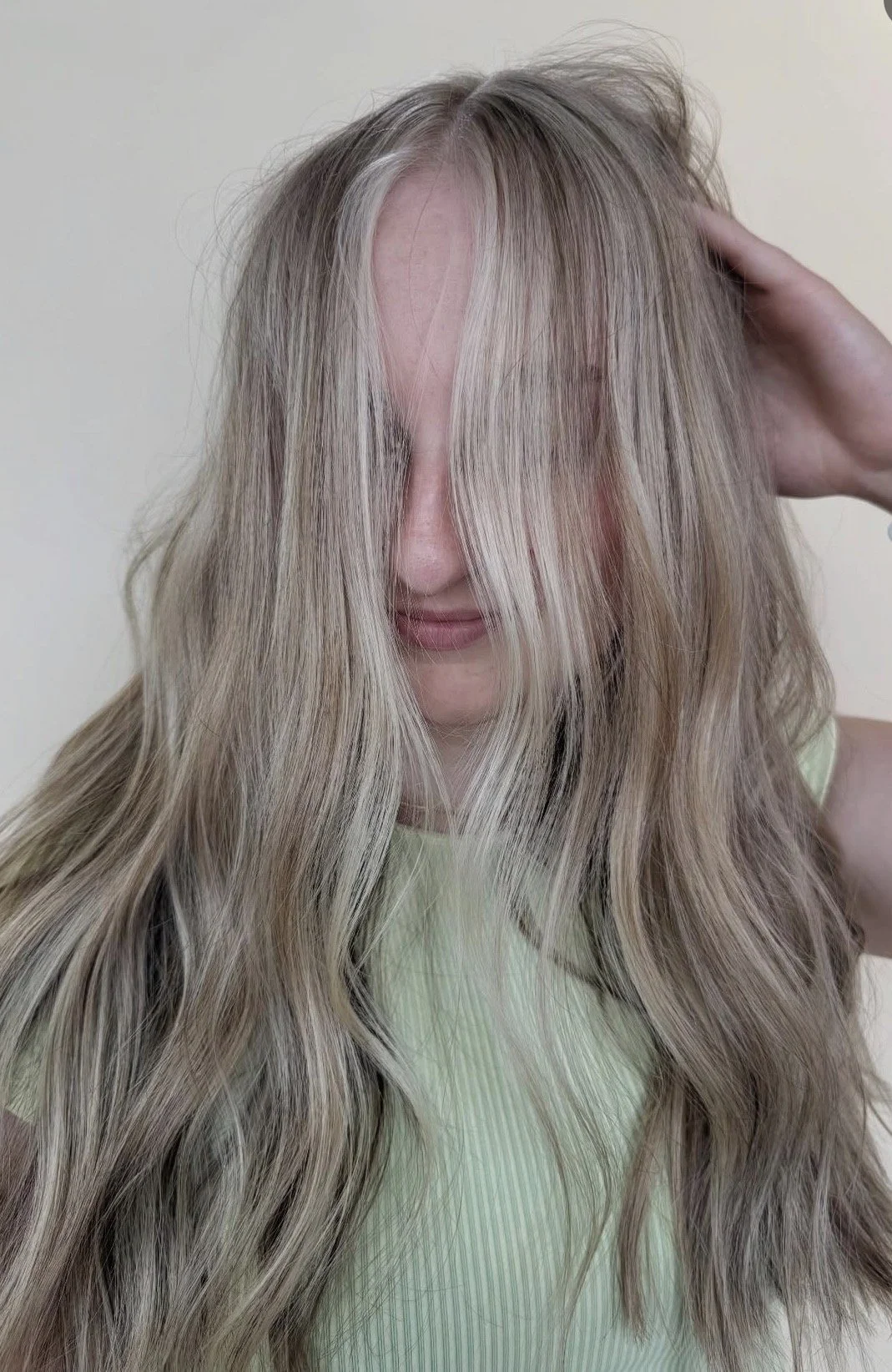 Person with long, wavy blonde hair covering face, raising hand to head, wearing a light-colored top, against a plain wall background.