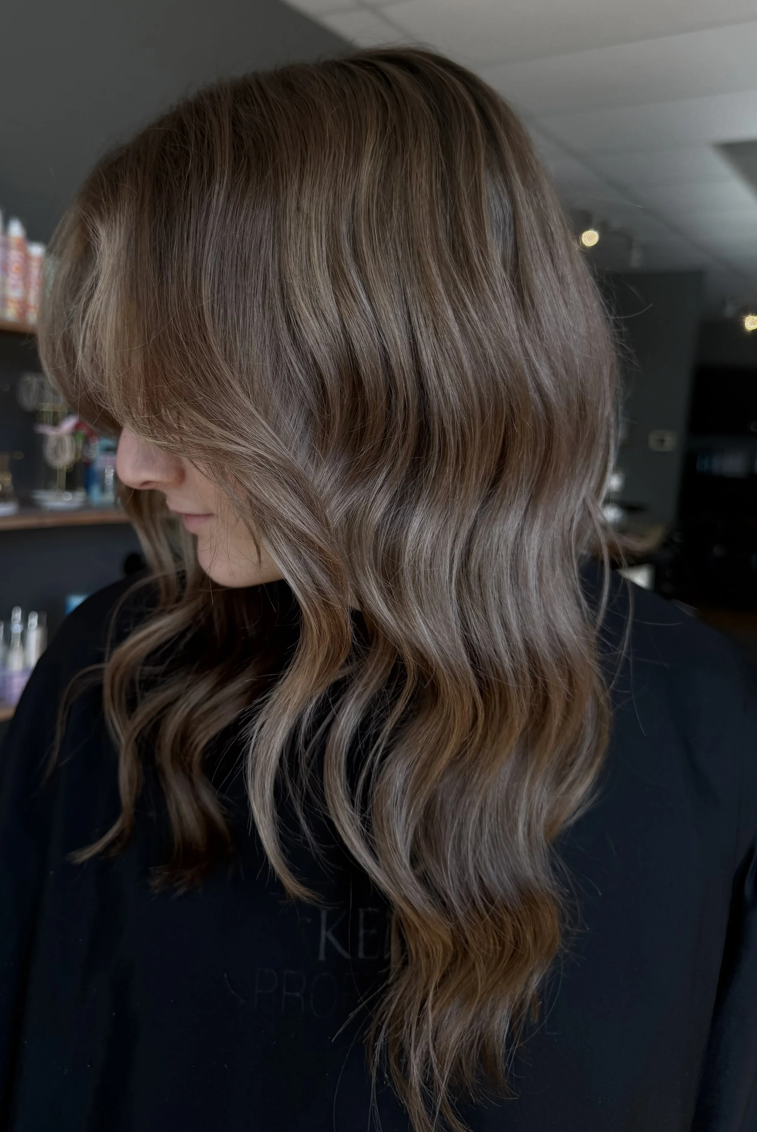 A woman with wavy, light brown hair styled in loose waves, looking to the side, in a hair salon.