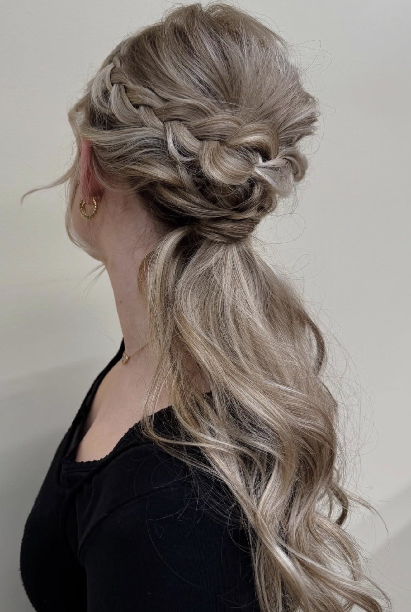 A woman with styled blonde hair in loose curls, featuring an intricate braided updo at the crown, wearing gold hoop earrings and a black top.