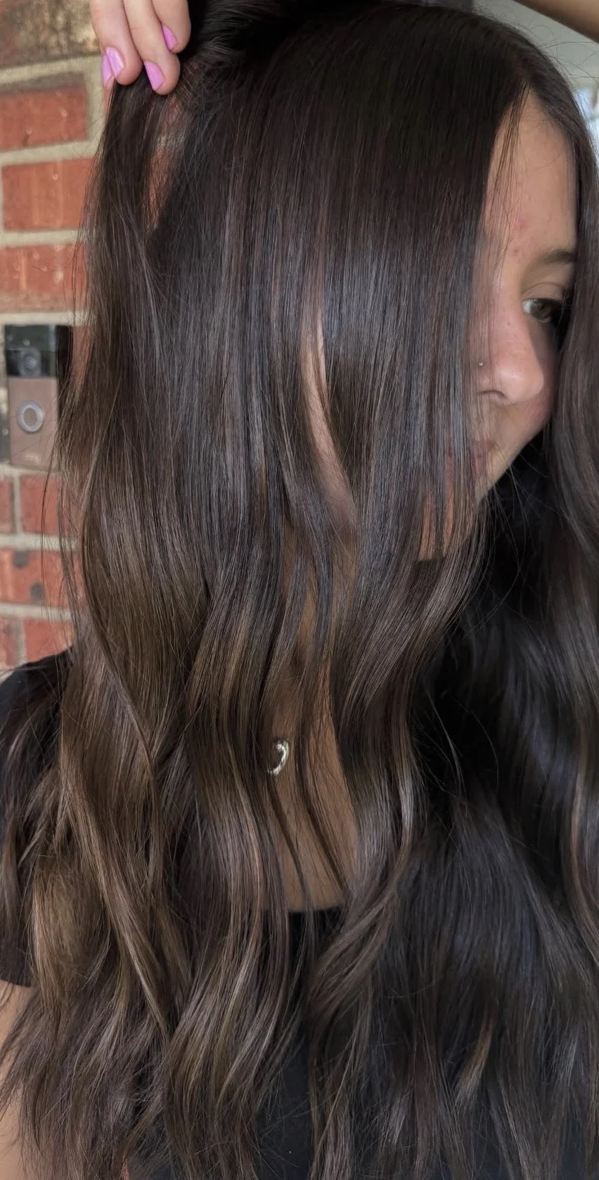 Close-up of a woman with long, wavy, dark brown hair with lighter brown highlights, partially covering her face, standing in front of a brick wall.