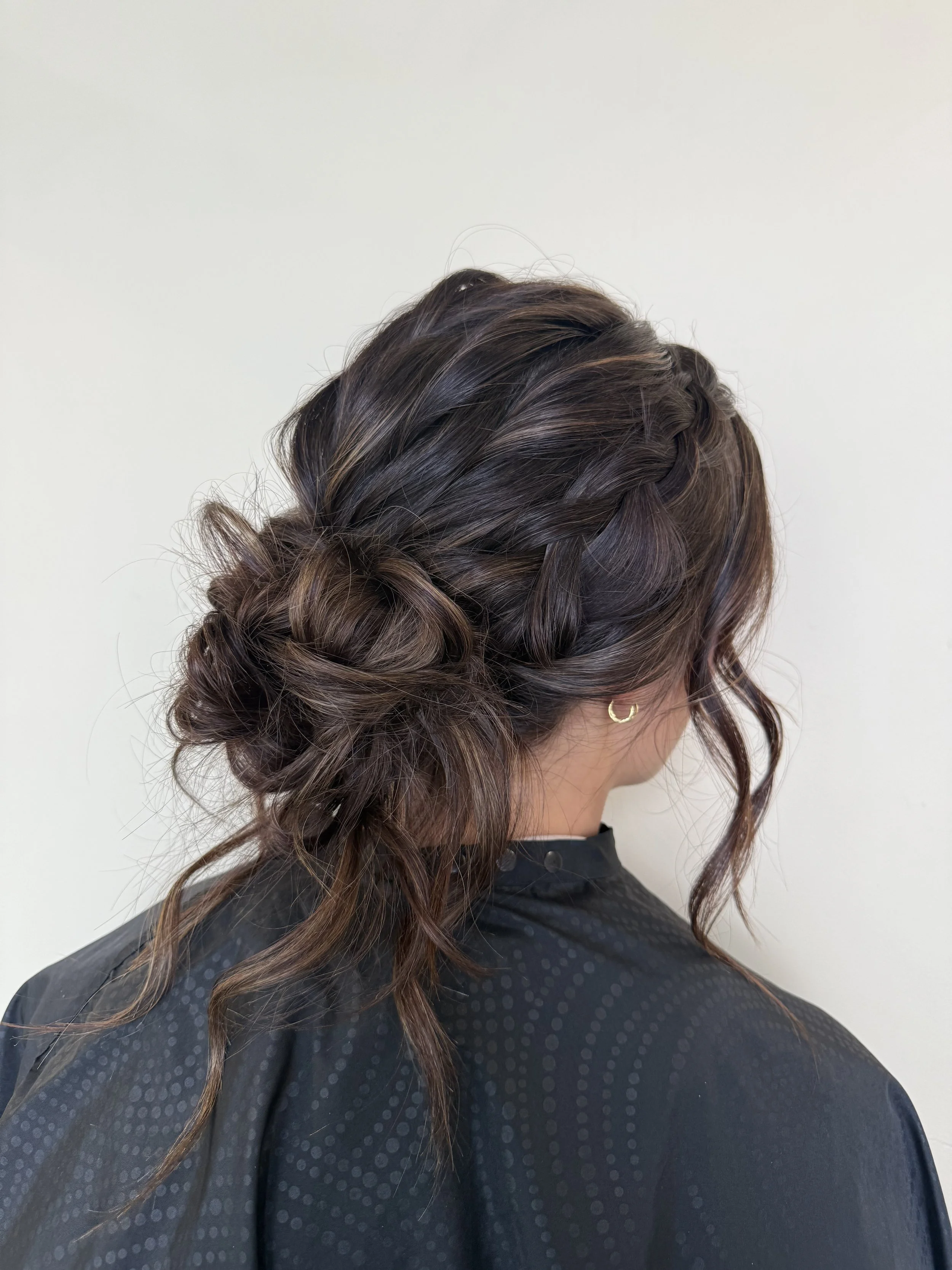 Back view of a woman with an elegant, loose braided updo hairstyle with waves and curls, wearing a black polka-dot top and gold hoop earrings.