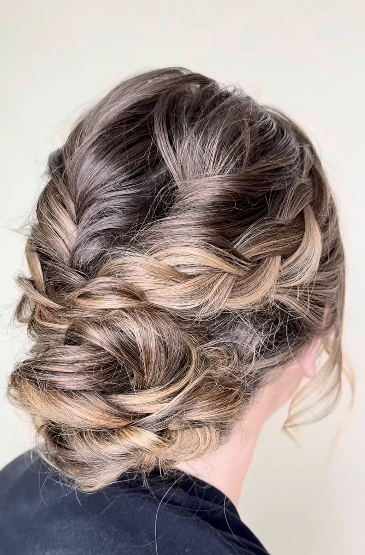 A close-up of a woman's styled hair in an intricate braid and bun, with light brown and blonde highlights.