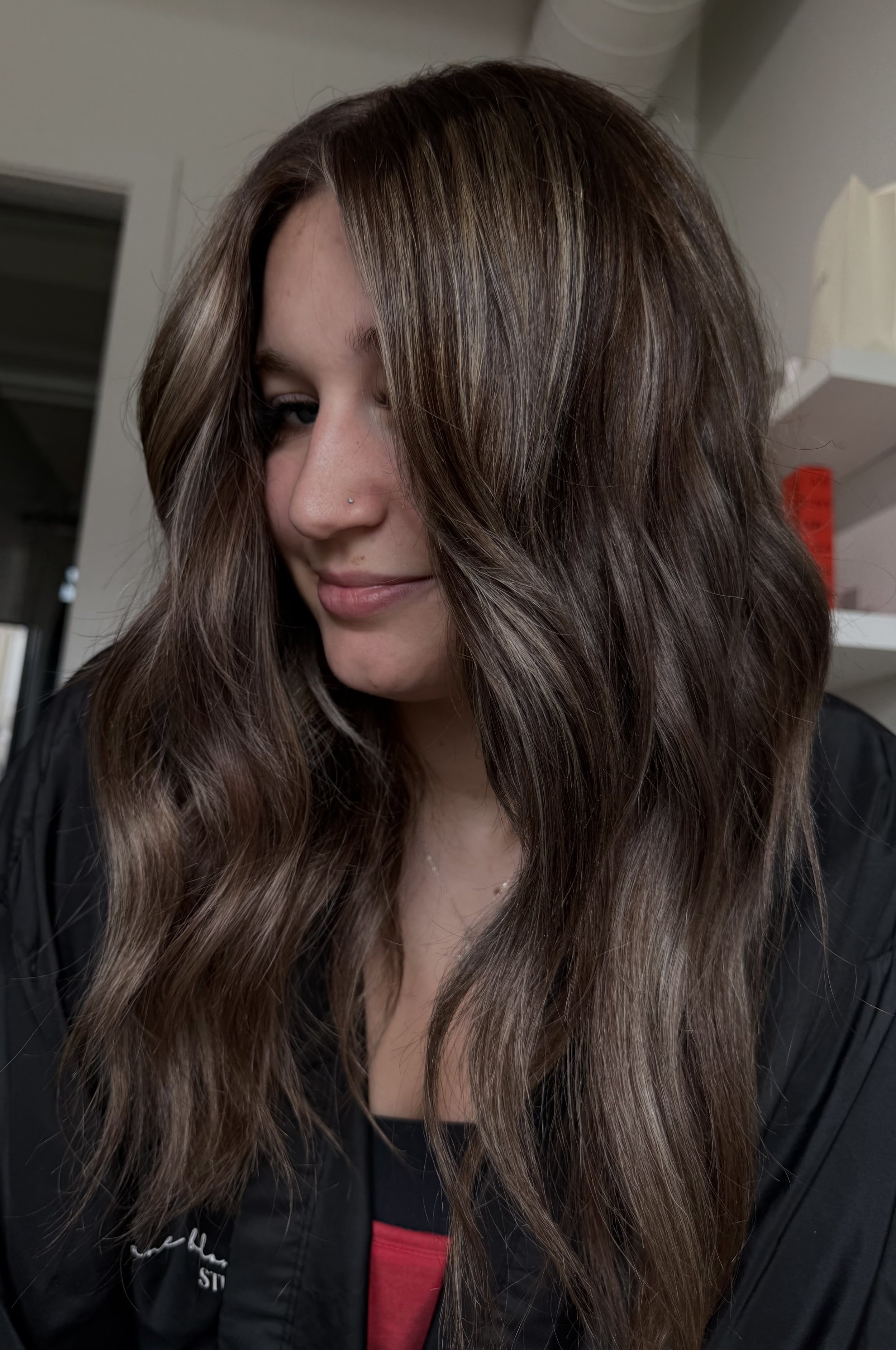 A young woman with long, wavy brown hair tilts her head slightly downward and to the side, with part of her face visible, showing a small nose piercing.