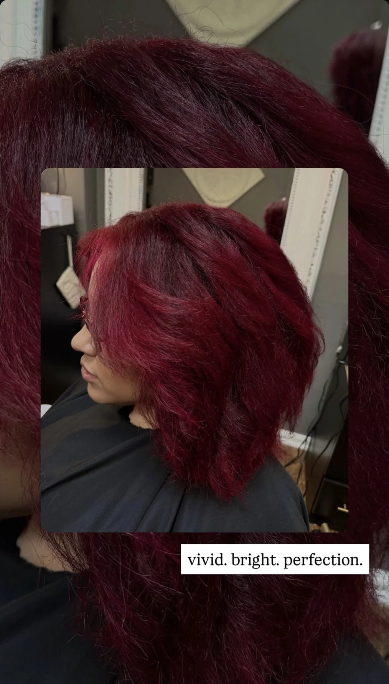 A woman with vibrant, bright red hair sitting in a salon chair, with a mirror and salon equipment in the background.