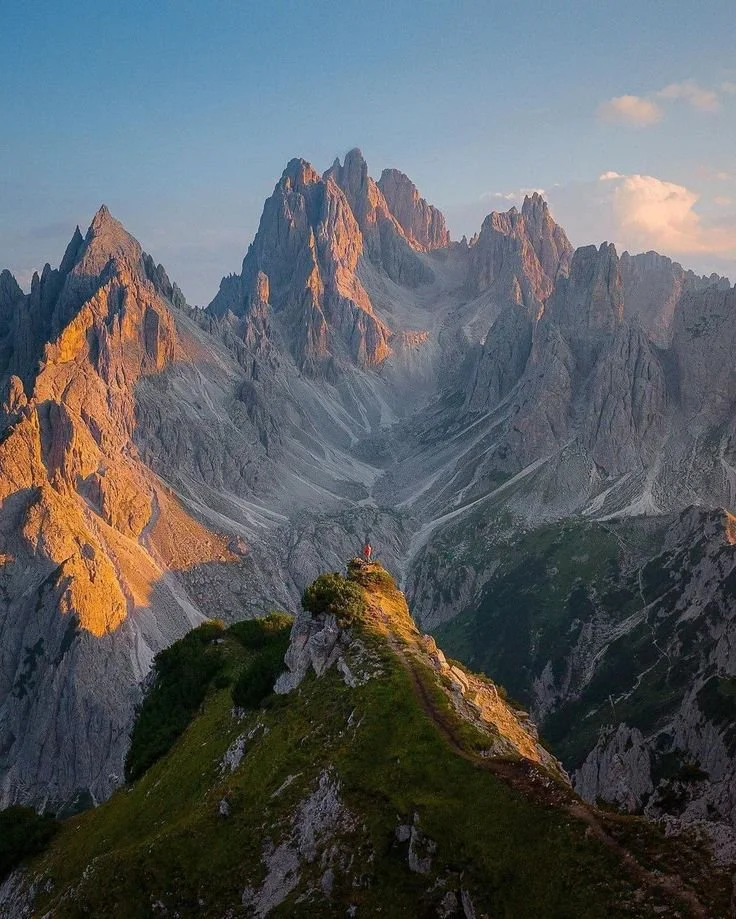 A mountain landscape featuring rugged peaks with warm sunlight, a green grassy trail ridge, and a person standing at the edge.