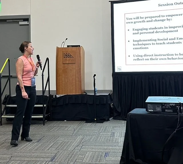 A woman holding a microphone stands in front of a presentation screen and podium during a conference in a hotel meeting room.