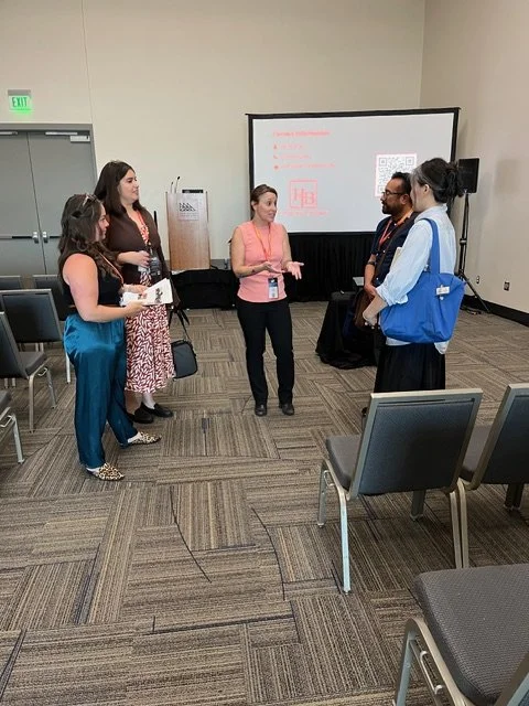 Five people in a conference room engaged in a discussion, with a presentation screen in the background displaying text and QR code, and black chairs arranged around the room.