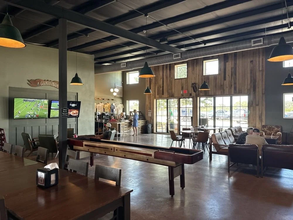 A spacious restaurant with wooden walls and large windows, featuring various seating areas including bar stools, sofas, and tables. A TV is mounted on the wall playing a football game, and a foosball table is in the foreground. People are sitting and walking inside, with natural light coming through the windows.