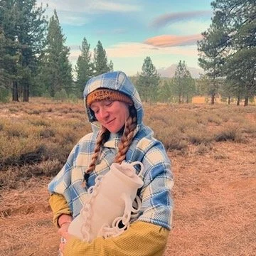 A young girl with braided hair standing outdoors in a forested area, wearing a blue plaid hoodie and holding a pair of white roller skates.
