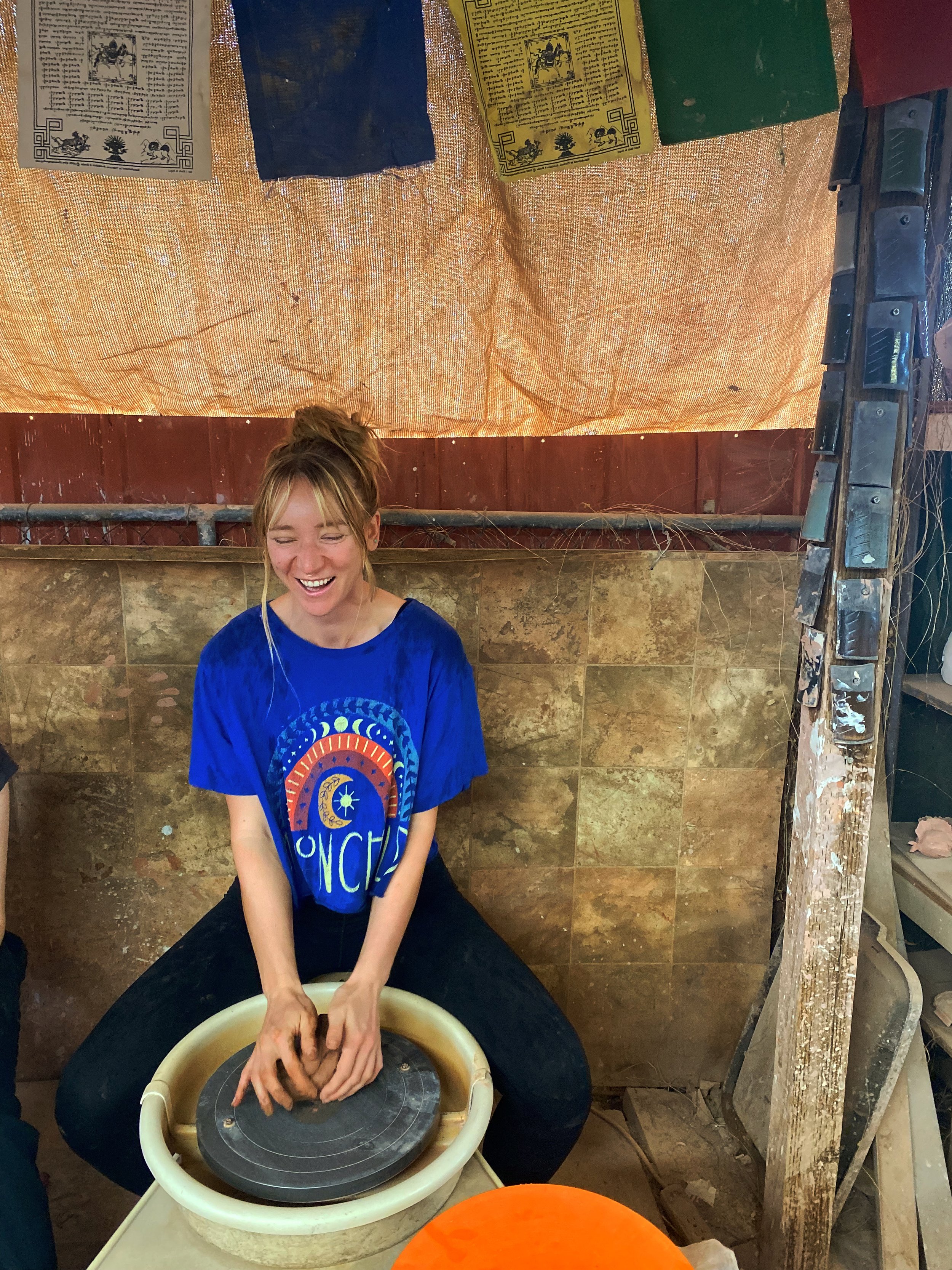 A woman with light hair tied in a bun, wearing a bright blue t-shirt with colorful patterns, is sitting on a stool and shaping clay on a pottery wheel, smiling and appearing happy.