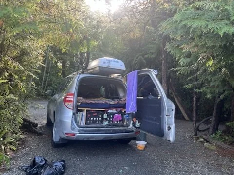 A silver hatchback car parked on a dirt path surrounded by trees, with its rear hatch open and a purple towel hanging from the door.