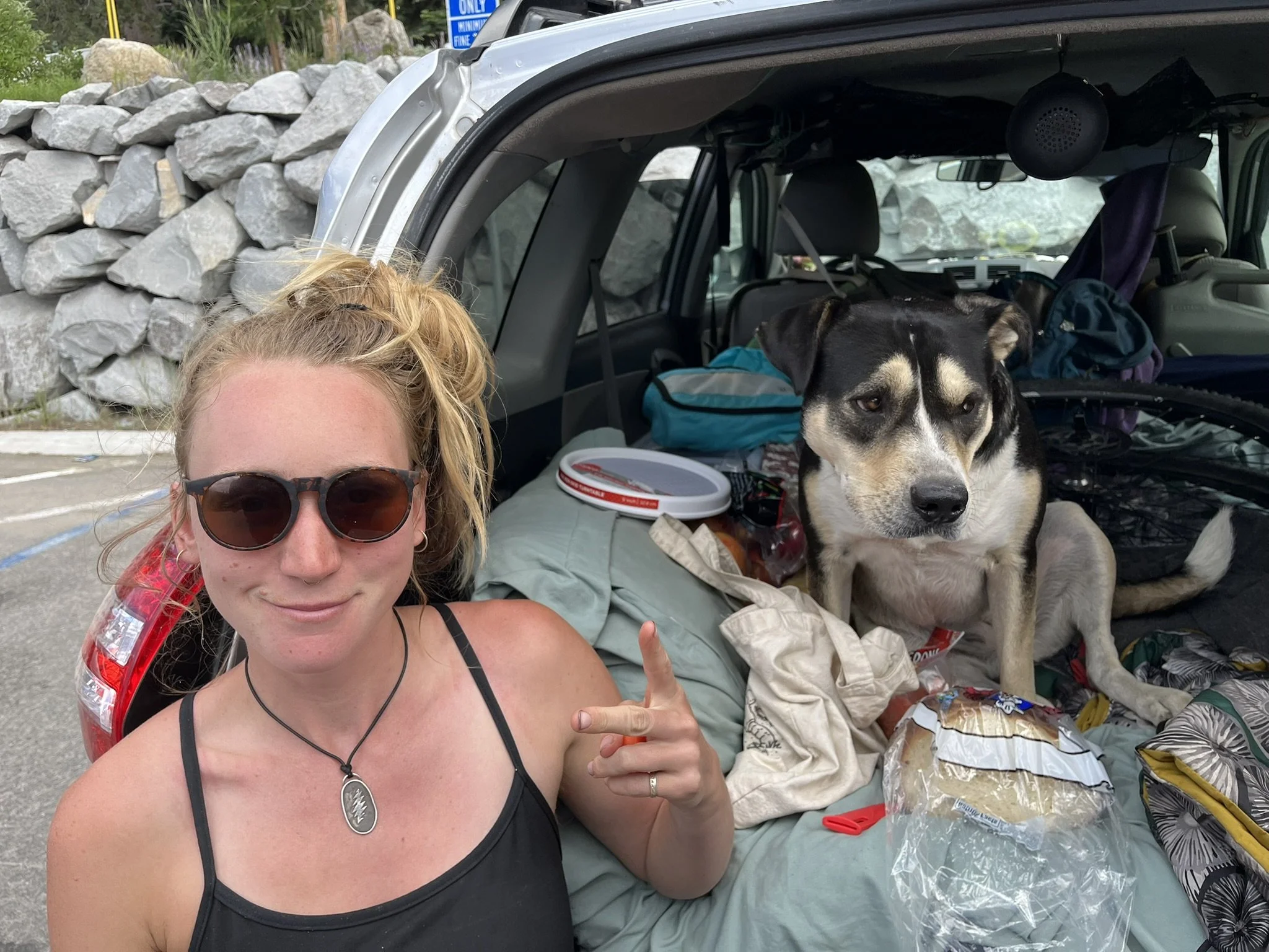 A woman with sunglasses and a black tank top smiling and making a peace sign next to a dog sitting inside an open car trunk filled with clothes and supplies.