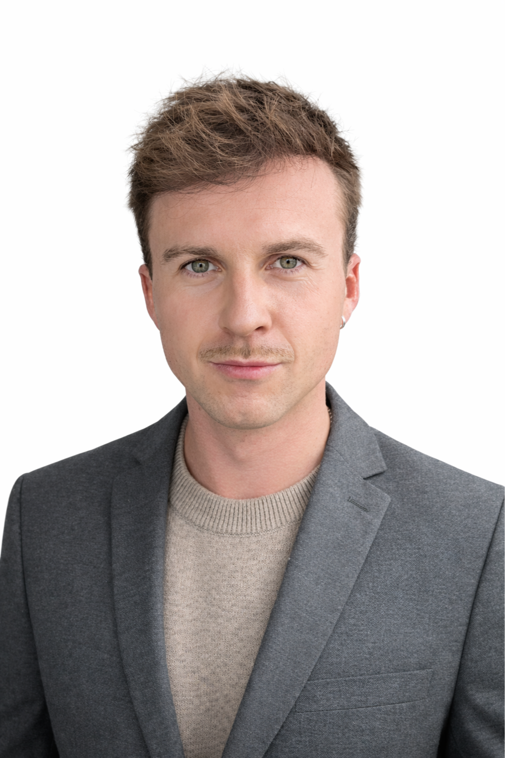 Un homme jeune avec des cheveux courts, portant un blazer gris et un pull beige, regardant l'objectif avec un visage sérieux devant un fond blanc.