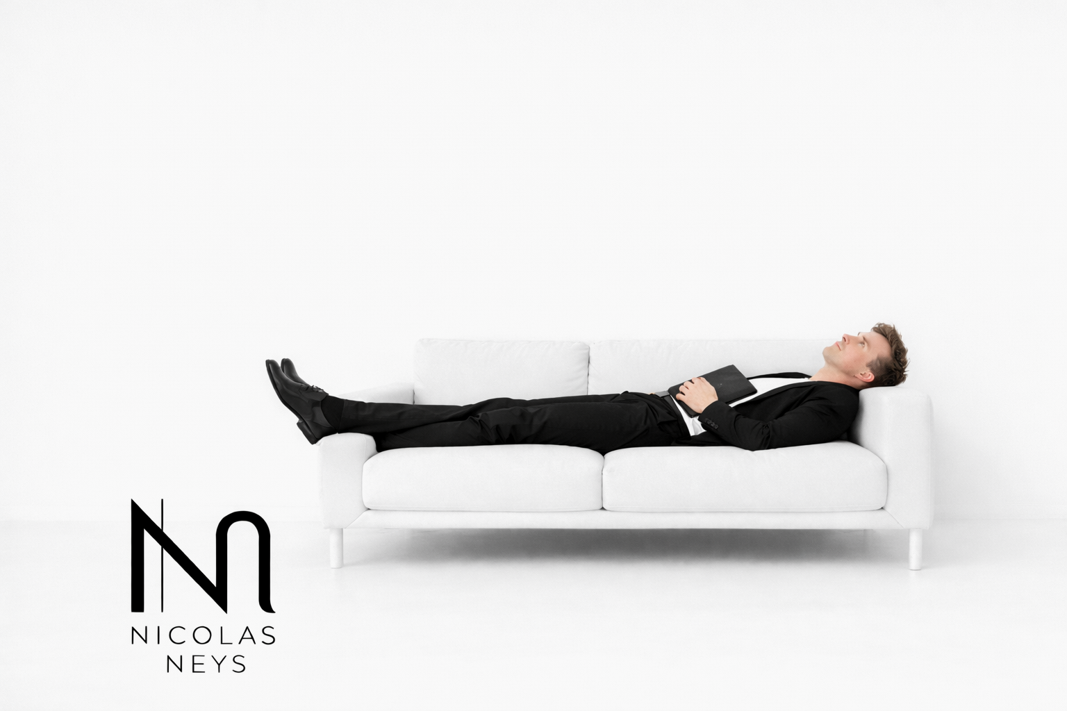 Homme allongé sur un canapé blanc, portant un costume noir, tenant un livre, dans un décor minimaliste blanc. Logo "Nicolas Neys" en bas à gauche.
