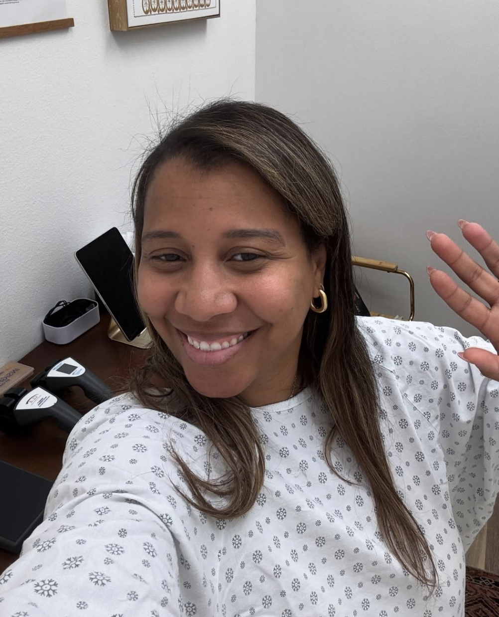A woman smiling and waving in a medical gown, taking a selfie in a room with medical equipment on a desk.