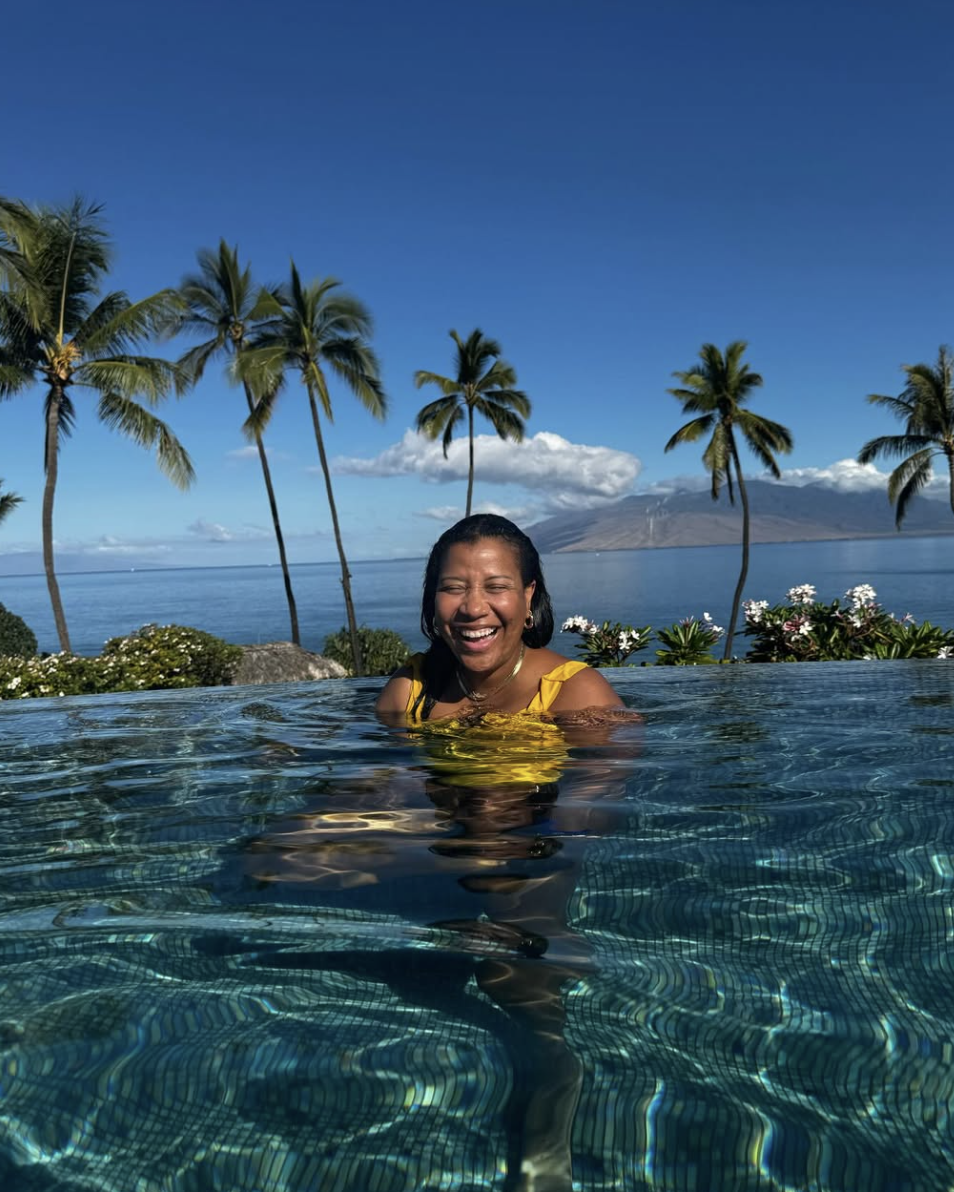 Woman smiling and enjoying herself in an infinity pool with a tropical landscape of palm trees, ocean, and mountains in the background under a clear blue sky.