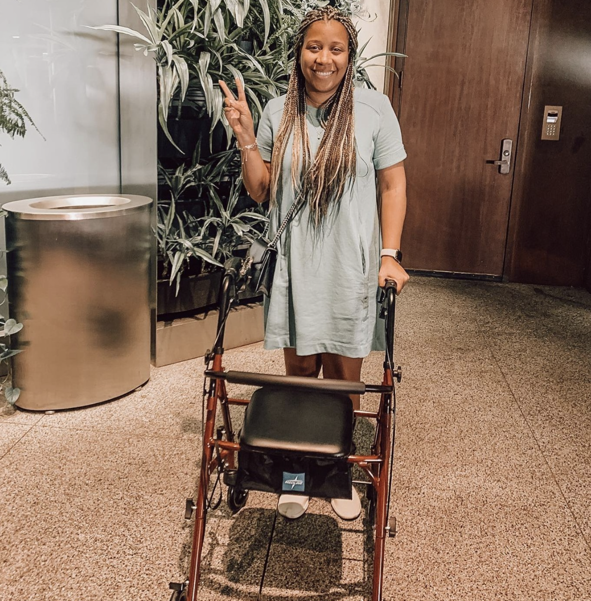 A woman standing indoors, smiling, making a peace sign with her right hand, with a walker in front of her, wearing a light gray dress and carrying a black purse