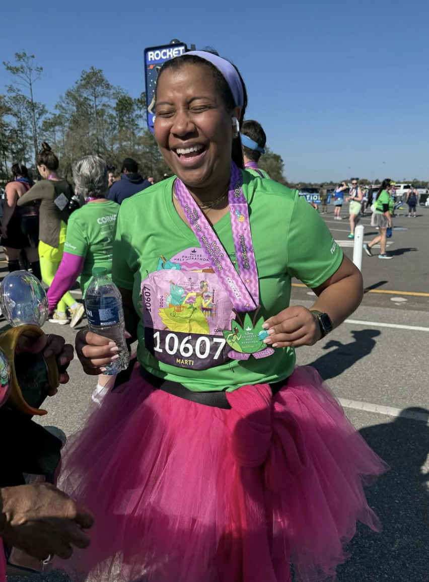 A woman wearing a green race shirt, pink tutu, and medal, smiling and holding water at a marathon event with other participants in the background.