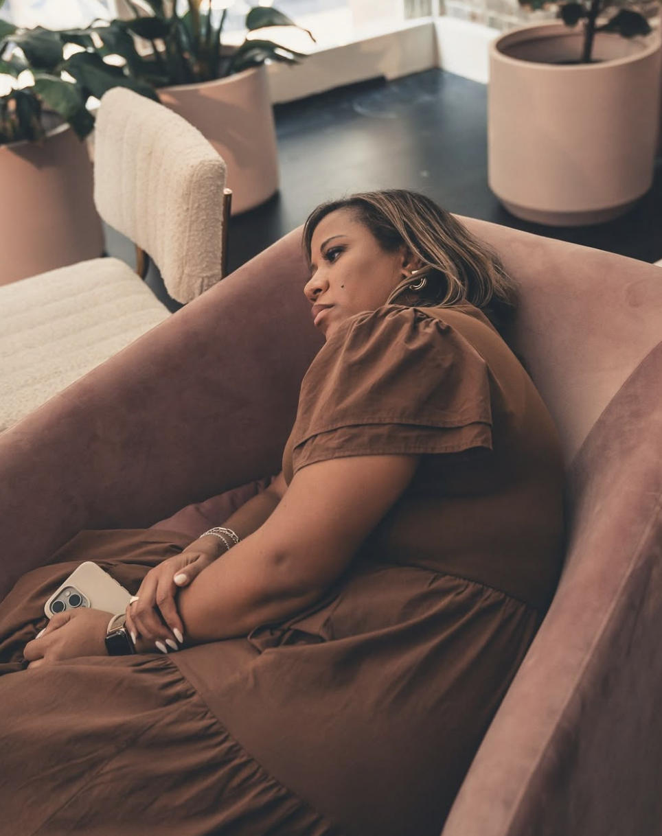 A woman is lying on a pinkish couch in a modern indoor space, holding a cellphone in her hand. She appears to be resting or lost in thought, with a neutral expression. The background features large potted plants and furniture.