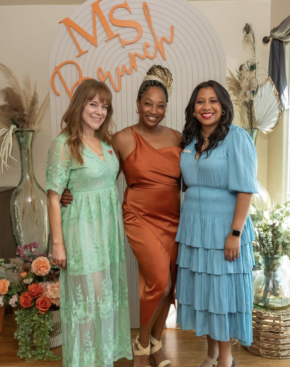 Three women standing together indoors in front of a decorative backdrop with the words 'MS Dream' written in orange. They are smiling and dressed in colorful dresses, with flowers and decorative dried plants around them.
