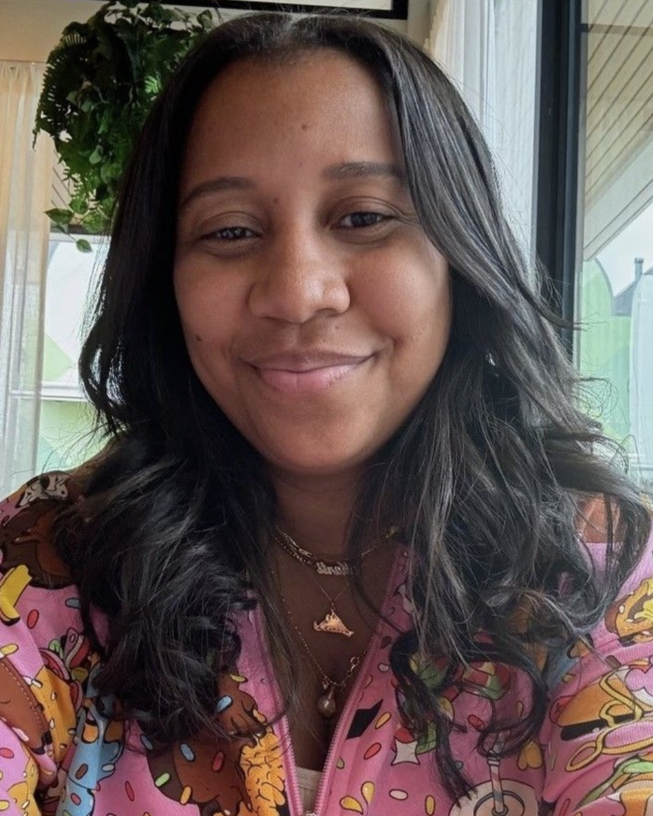 A woman with black wavy hair smiling, wearing a colorful pink jacket with cartoon and food print, adorned with layered necklaces, sitting in front of a window with curtains and greenery outside.