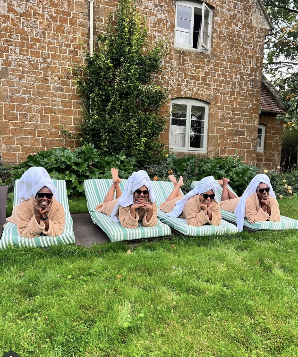 Four people lying on striped lounge chairs outdoors, wearing beige robes and white towels on their heads, smiling and wearing sunglasses, in a garden with green grass and leafy plants in front of a brick house.
